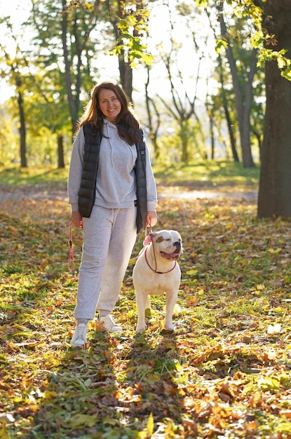 犬は公園で愛人と遊ぶ ジャケットを着た女性と、公園の黄色い紅葉の中で遊ぶアメリカン ブルドッグ犬のクローズ アップ