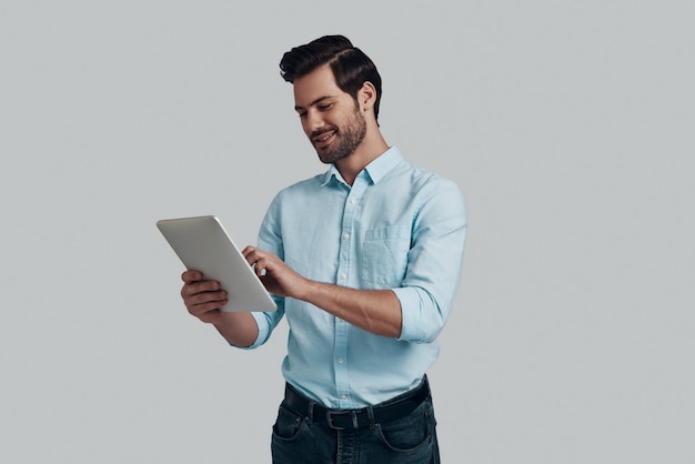 Doing business. Handsome young man working using digital tablet while standing against grey background