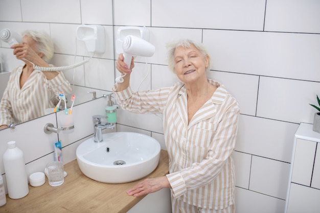 Doing hair. Gray-haired aged woman drying her hair in the bathroom