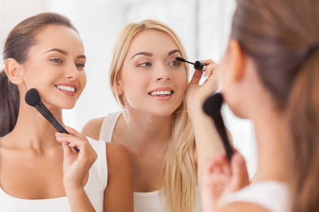 Doing make-up together. Two beautiful young women doing make-up together while looking at the mirror and smiling