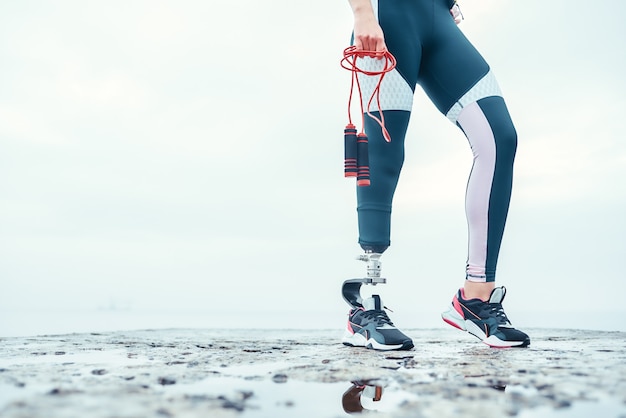 Фото Я стараюсь изо всех сил. обрезанное изображение женщины-инвалида в спортивной одежде с протезом ноги, держащей скакалку, стоя перед морем. спортсмен-инвалид. здоровый образ жизни. кардио