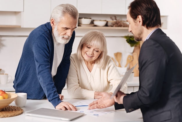 Doing my job properly . Skillful optimistic sincere real estate agent having conversation with elderly customers and using important documents while representing the house