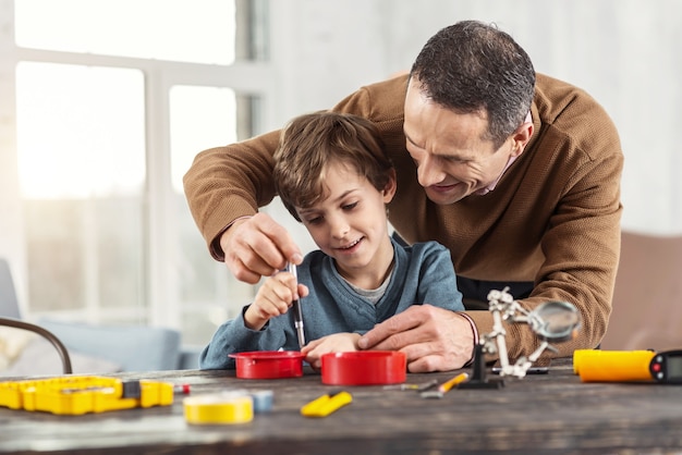 Doing things together. Handsome alert little fair-haired boy smiling and learning how to use instruments and his father standing behind him and helping him