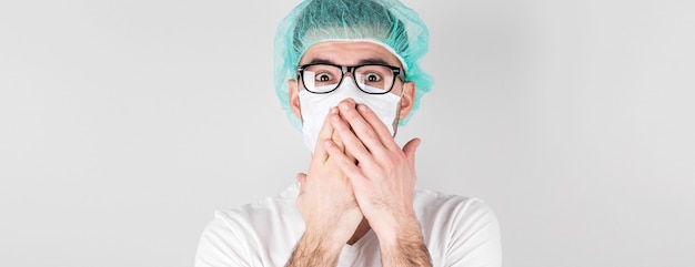 Dokter-chirurg in wit medisch masker en een medische pet staat met verbazing op witte achtergrond, heeft betrekking op zijn mond met de handen. Ð¡ concept van covid 19, griep en seizoensgebonden verkoudheid.