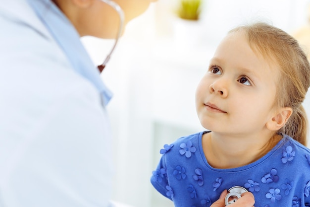 Dokter die een kind onderzoekt met een stethoscoop in een zonnige kliniek. gelukkig lachend meisje patiënt gekleed in blauwe jurk is bij de gebruikelijke medische keuring.