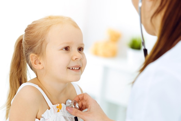 Dokter die een kind onderzoekt met een stethoscoop in een zonnige kliniek. Gelukkig lachend meisje patiënt gekleed in felle kleuren jurk is bij de gebruikelijke medische keuring.