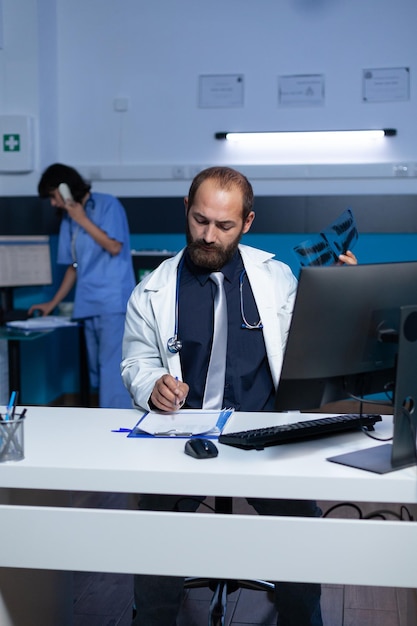 Dokter die radiografie vasthoudt voor onderzoek terwijl hij aantekeningen maakt op het controledocument. Dokter schrijft diagnose en resultaten na röntgenscananalyse voor medische behandeling om middernacht.