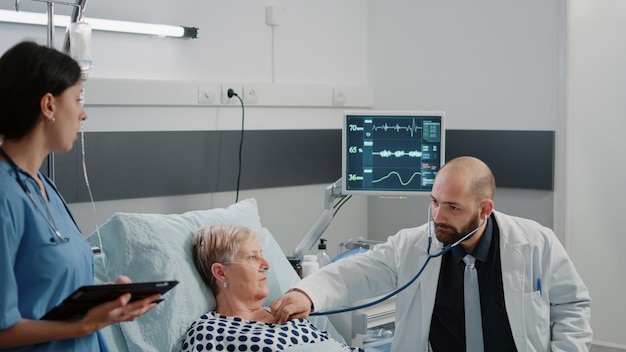 Dokter die stethoscoop gebruikt voor hartslagmeting bij zieke patiënt in ziekenhuisbed. Dokter doet gezondheidszorgoverleg met bejaarde vrouw terwijl verpleegster met tablet hulp geeft