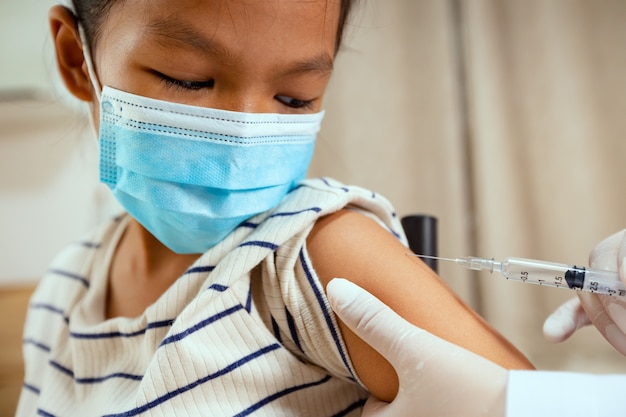 Dokter die vaccinatie op de aziatische arm van het kindmeisje injecteert. Het kindmeisje dat een beschermend gezichtsmasker draagt om de pandemie van het Coronavirus Covid-19 te beschermen. Gezondheidszorg en medisch concept.