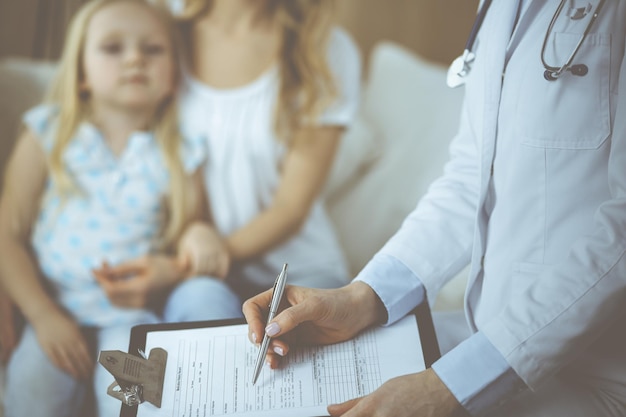 Dokter en patiënt. Kinderarts die klembord gebruikt terwijl hij een klein meisje met haar moeder thuis onderzoekt. Ziek en ongelukkig kind bij medisch onderzoek. Geneeskunde concept.