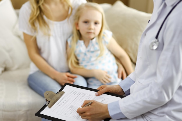 Dokter en patiënt. Kinderarts die klembord gebruikt terwijl hij een klein meisje met haar moeder thuis onderzoekt. Ziek en ongelukkig kind bij medisch onderzoek. Geneeskunde concept.