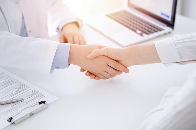 Dokter en patiënt schudden elkaar de hand terwijl ze tegenover elkaar aan tafel zitten in de kliniek, alleen de handen van dichtbij. Geneeskunde concept.
