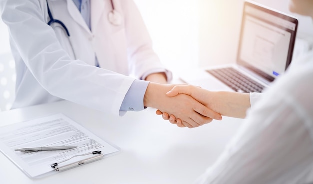 Dokter en patiënt schudden elkaar de hand terwijl ze tegenover elkaar aan tafel zitten in de kliniek, alleen de handen van dichtbij. Geneeskunde concept.