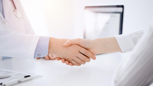 Dokter en patiënt schudden elkaar de hand terwijl ze tegenover elkaar aan tafel zitten in de kliniek, alleen handen van dichtbij. Geneeskunde concept