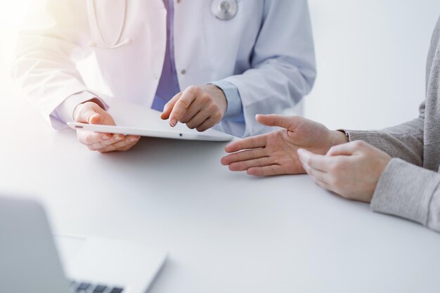 Dokter en patiënt zitten aan de tafel in de kliniek. De focus ligt op de handen van de vrouwelijke arts met behulp van een tabletcomputer, close-up. Geneeskunde en gezondheidszorgconcept