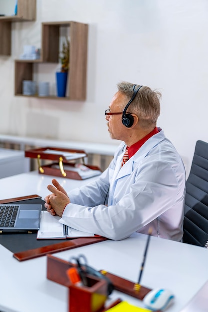 Dokter geeft consultatie in medische kamer via laptop Ervaren medic in scrubs zit aan tafel in koptelefoon Closeup