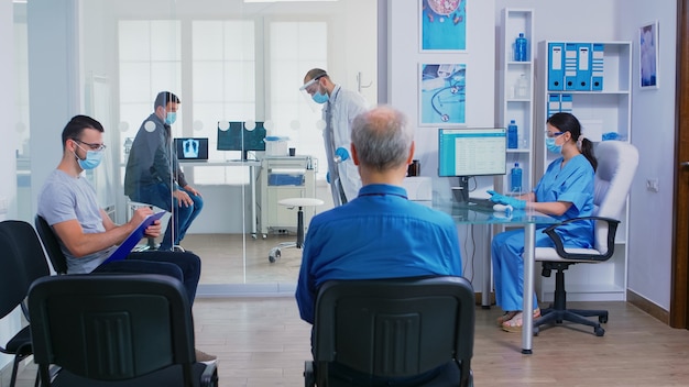 Dokter geeft verpleegster patiënt radiografie terwijl hij het onderzoek van de zieke man in zijn kantoor afrondt. Assistent met gezichtsmasker tegen covid-19 die gehandicapte vrouw in rolstoel door wachtruimte duwt.