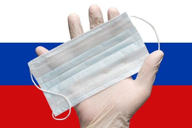 Dokter houdt gezichtsmasker in de hand in witte medische handschoen op achtergrondkleuren vlag van Rusland. Concept pandemische verzekering coronavirus, griep, virusquarantaine, pandemische uitbraak, grippe, door de lucht overgedragen ziekten