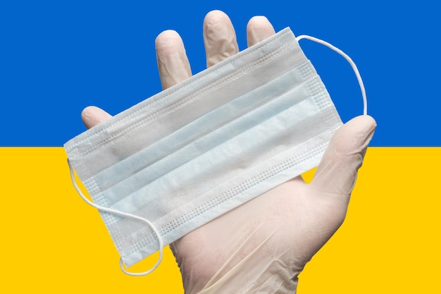 Dokter houdt medisch gezichtsmasker in de hand in witte handschoenen op achtergrondkleuren Nationale vlag van Oekraïne Concept coronavirusziekte SARSCoV2 pandemisch virus quarantaine-uitbraak grippe