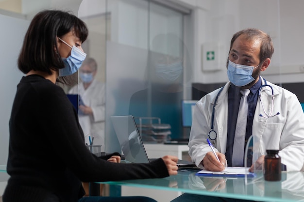 Dokter in overleg met vrouw die aantekeningen maakt op klembordpapier, ontmoeting met volwassene om medisch advies te geven. Man die een diagnose van de gezondheidszorg schrijft tijdens een controlebezoek tijdens de covid 19-pandemie.