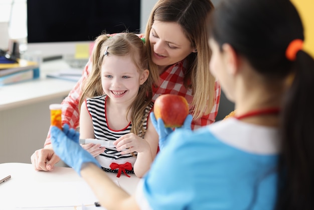 Dokter kinderarts met fles met medische capsules en rode appel voor klein meisje en m...