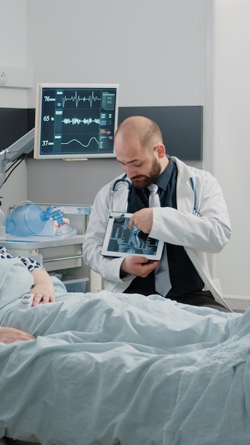 Dokter legt röntgenscanresultaten op tablet uit aan bejaarde patiënt in de faciliteit. Dokter die de radiografiediagnose op het apparaatscherm analyseert voor gezondheidszorg en herstelbehandeling op de ziekenhuisafdeling.