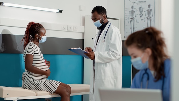 Dokter maakt aantekeningen bij onderzoek met zwangere vrouw tijdens covid 19 pandemie in medische kast. mannelijke verloskundige die patiënt raadpleegt die een kind verwacht en controledocumenten gebruikt.