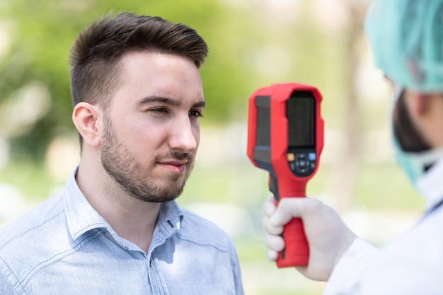 Dokter Man gebruik infrarood voorhoofd thermometer pistool om de lichaamstemperatuur te controleren op virus Covid-19 symptomen met de isolatiejurk of beschermende pakken en chirurgische gezichtsmaskers buiten in het park