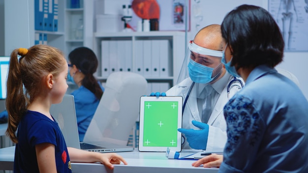 Dokter met behulp van groen scherm tablet in medisch kantoor tijdens covid-19. Gezondheidszorgspecialist met chroma key notebook geïsoleerd mockup vervangingsscherm. Eenvoudig medisch medisch gerelateerd thema intoetsen.