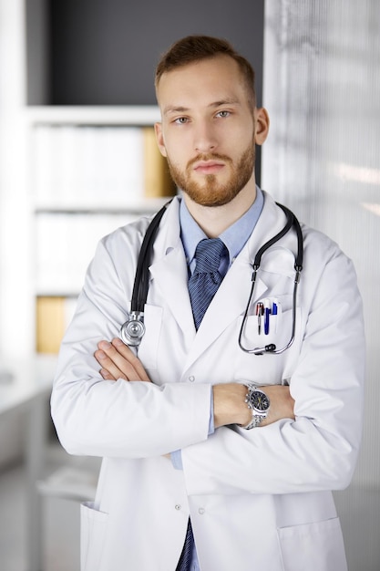 Dokter met rode baard die rechtop in de kliniek staat, vlakbij zijn werkplek. Portret van arts. Geneeskunde, gezondheidszorg