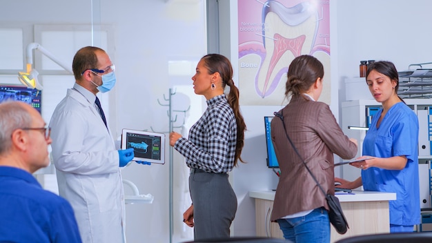 Dokter met tablet met röntgenfoto die het aan de patiënt laat zien terwijl de verpleegster de vrouw helpt bij het invullen van een tandheelkundig document. Stomatoloog presenteert tandheelkundige radiografie met behulp van moderne gadget in stomatologische kliniek