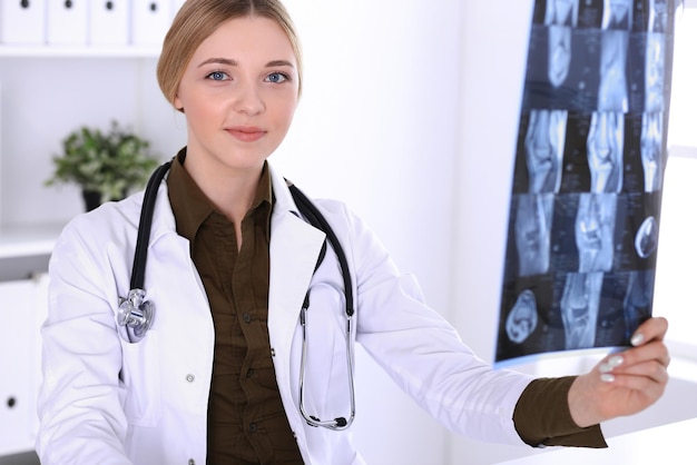 Dokter vrouw onderzoekt x-ray foto in de buurt van raam in het ziekenhuis. Chirurg of orthopedist op het werk. Geneeskunde en gezondheidszorg concept. Kakikleurige blouse van een therapeut ziet er goed uit.