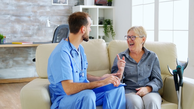 Dokterassistent die aantekeningen maakt op het klembord terwijl hij luistert naar oude gepensioneerde vrouw in verpleeghuis. Verzorger en maatschappelijk werker