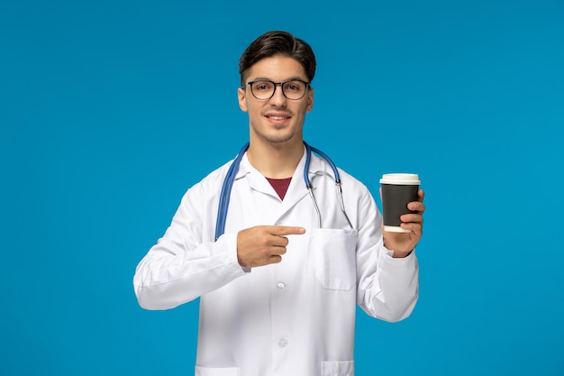 Doktersdag knappe brunette schattige kerel in medische jurk wijzend op koffiekopje met bril