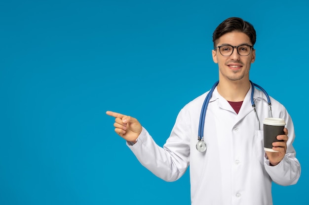 Doktersdag schattige jonge brunette man in laboratoriumjas die een bril draagt en glimlacht en naar links wijst