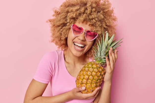 Dolblij krullende vrouw lacht vreugdevol houdt verse ananas gaat maken van vers sap van het poses met favoriete exotisch fruit terloops gekleed geïsoleerd op roze achtergrond Gezonde voeding