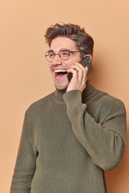 Dolblij man praat op mobiele telefoon heeft grappig gesprek bespreekt iets lacht vrolijk gekleed in vrijetijdskleding ergens geïsoleerd over beige achtergrond. Technologie concept