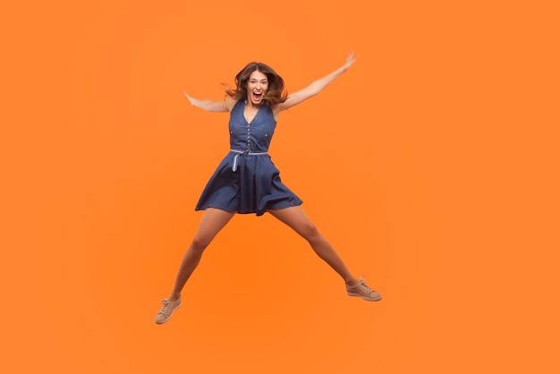 Dolblij opgewonden extatische brunette vrouw in denim jurk springen als een ster en schreeuwen van enthousiasme vliegen geïsoleerd op een oranje achtergrond vol gelukkig vreugdevolle emoties vrijheid en energie