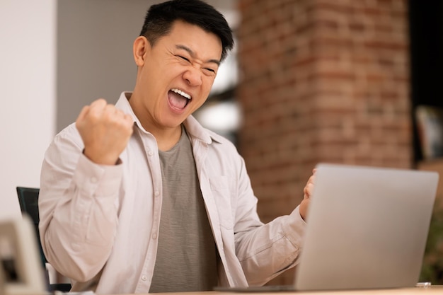 Dolblije aziatische man die laptop gebruikt en vuisten schudt om geweldig online nieuws en succes te vieren