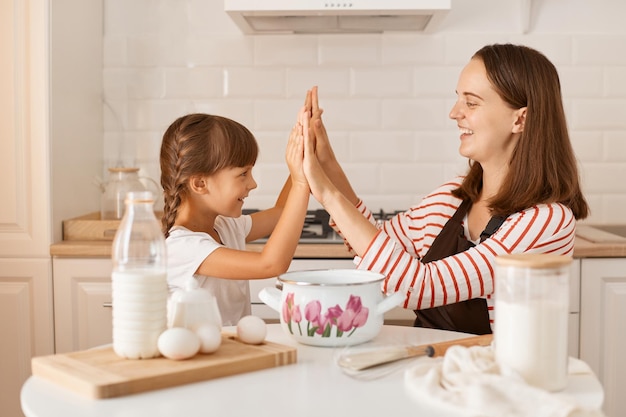 Dolblije donkerharige moeder veel plezier tijdens het bakken van koekjes of gebak met schattig klein kleutermeisje thuis, gelukkige jonge moeder leert koken samen met kleine dochter die high five geeft