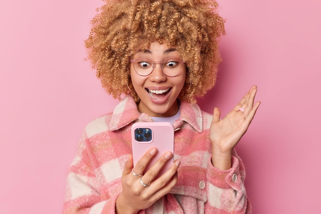 Dolblije jonge vrouw met krullend haar lacht aangenaam staart naar het scherm van de smartphone houdt de handpalm omhoog en voelt zich opgewonden door het winnen van grote kortingen in de online webwinkel controleert de meldingshoudingen binnen