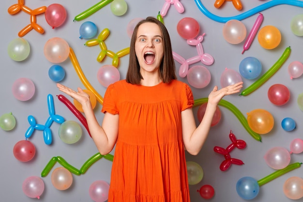 Dolblije vrouw Verraste emotie Opgewonden vrouw met bruin haar in een oranje jurk hief haar armen op terwijl ze schreeuwde wow staande tegen een grijze muur versierd met kleurrijke ballonnen