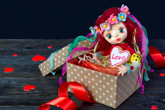 Doll sitting in a gift box with a heart. In the hands of a cookie