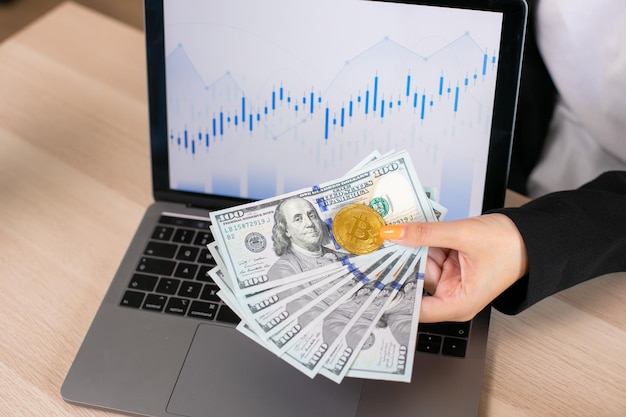 Photo dollar bills and bitcoin holding in hands on a background laptop with charts bitcoins growth chart