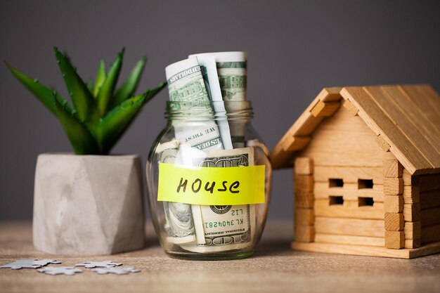 Dollar bills in glass jar and text house on wooden table.