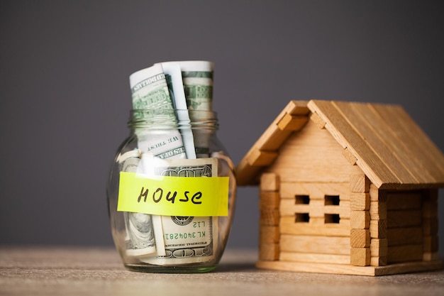 Dollar bills in glass jar and text house on wooden table.