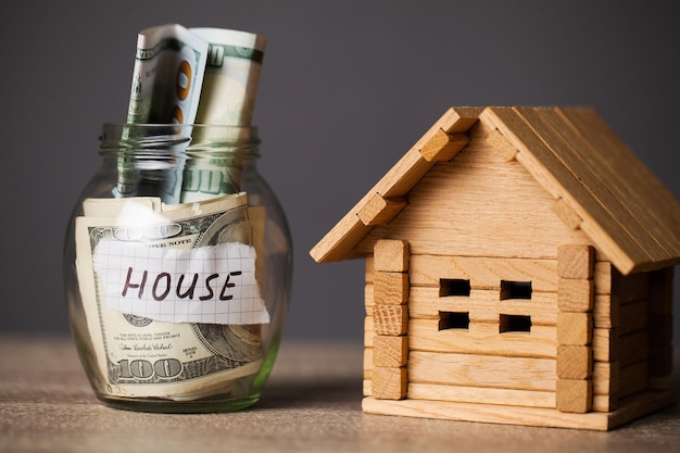 Dollar bills in glass jar and text house on wooden table.