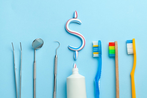 Dollar sign of toothpaste. Tube of colored toothpaste and a toothbrush and dentist tools, a mirror, a hook on a blue background. Concept expensive dentist services.