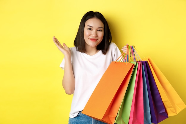 Dom Aziatisch meisje boodschappentassen tonen en schouderophalend, winkel in winkels met kortingen, staande over geel.