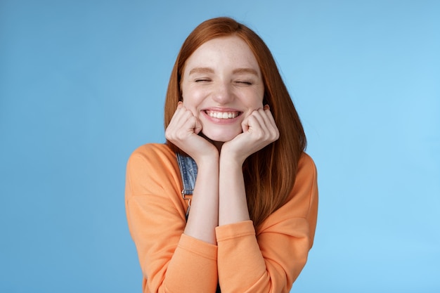 Dom schattig gelukkig roodharige vriendin glimlachend in het algemeen ogen dicht dromerig knijp wangen opgetogen gevraagd datum man houdt van staan blauwe achtergrond vreugde hebben fantastische gelukkige dag triomferen